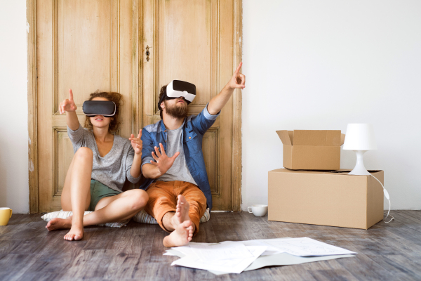 Young married couple moving in new house, sitting on the floor near cardboard boxes, wearing virtual reality goggles.