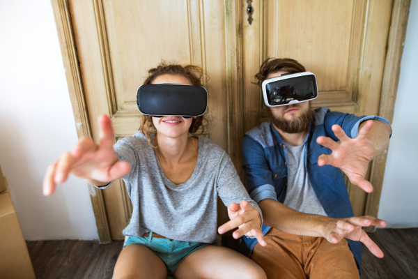 Young married couple moving in new house, sitting on the floor, wearing virtual reality goggles.