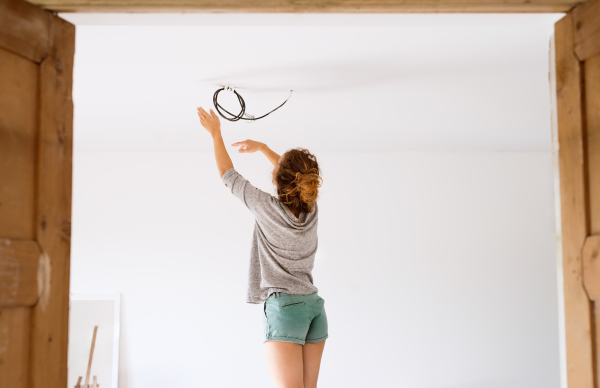 Beautiful young woman in a new house standing on stepladder, instaling a light. Rear view.