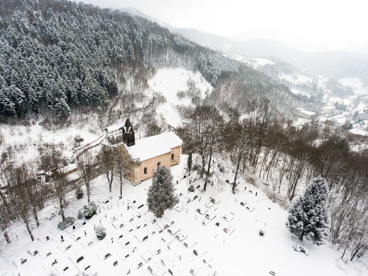 Aerial view of a church and a cemetery with hills and forest surrounding it. Village in winter time.