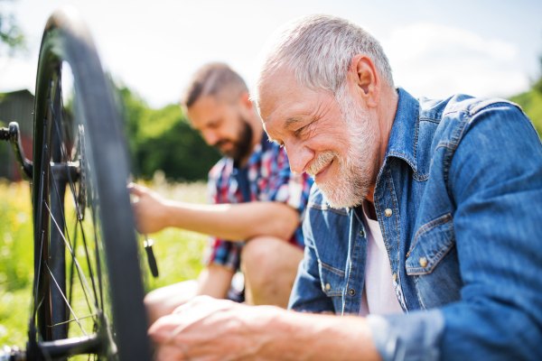 An adult hipster son and senior father repairing bicycle outside on a sunny summer day.