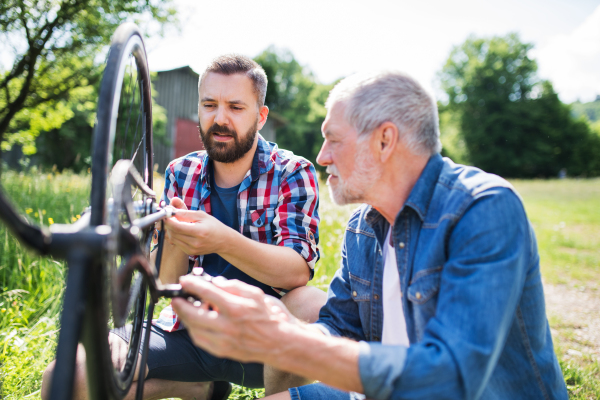 An adult hipster son and senior father repairing bicycle outside on a sunny summer day.