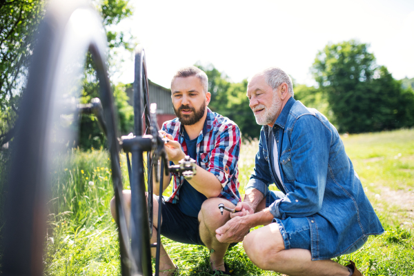 An adult hipster son and senior father repairing bicycle outside on a sunny summer day.