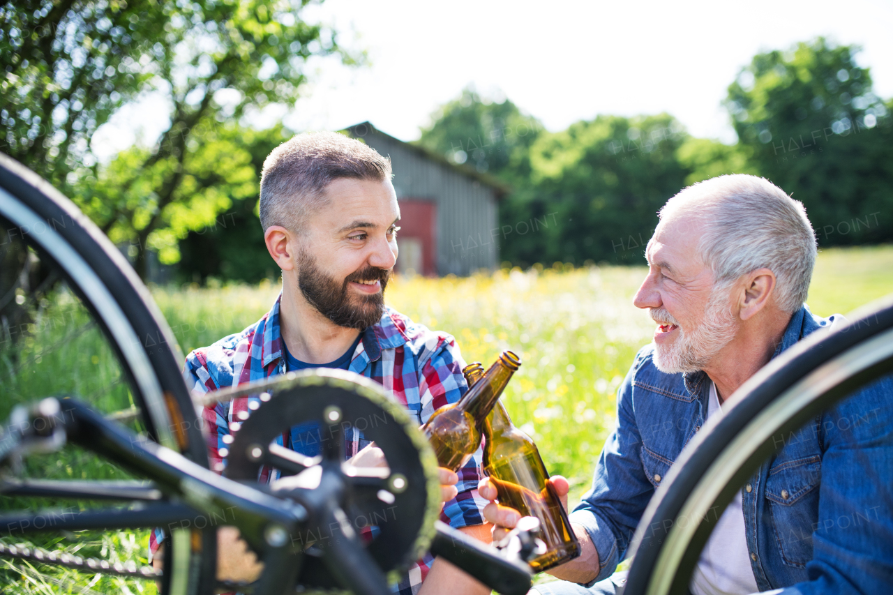 An adult hipster son and senior father repairing bicycle outside on a sunny summer day, drinking beer.