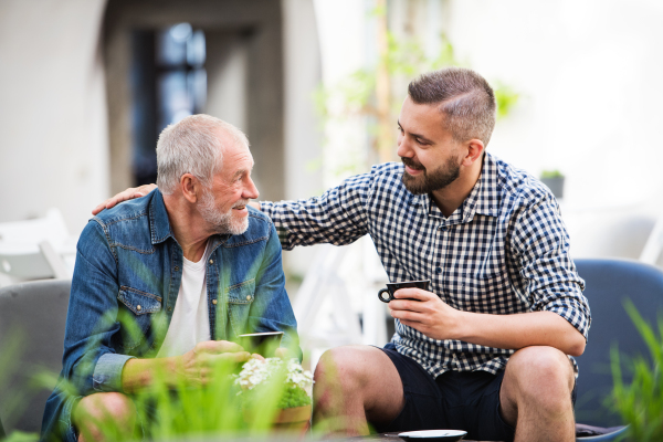 An adult hipster son and senior father with a cup of coffee in an outdoor cafe, talking.