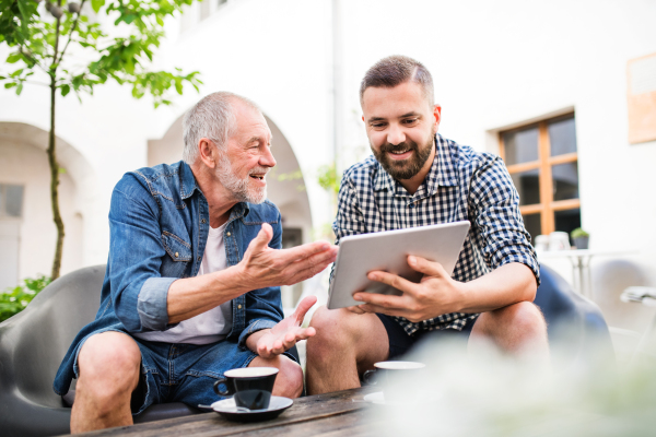 A happy adult hipster son with tablet and senior father in a cafe in town.