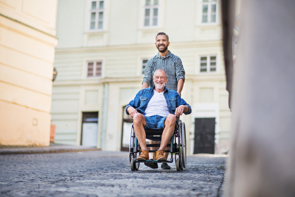 An adult hipster son with his senior father in wheelchair on a walk in town.