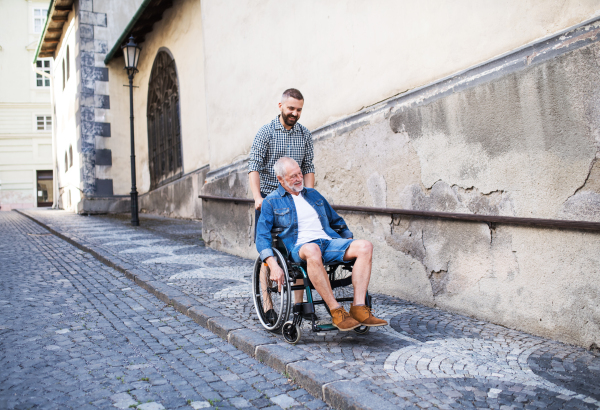 An adult hipster son with his senior father in wheelchair on a walk in town.