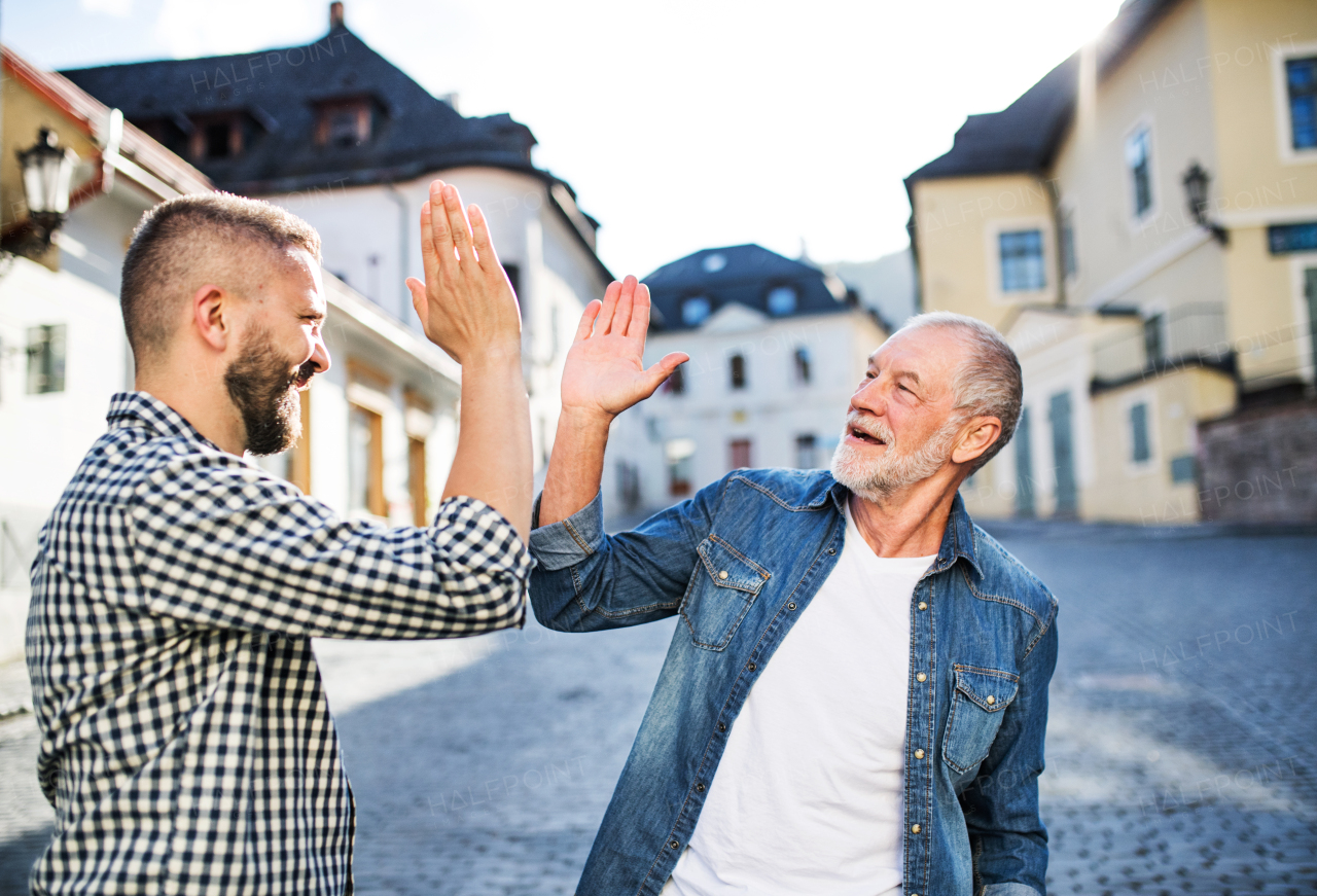 An adult hipster son and his senior father in town, giving high five when greeting each other.