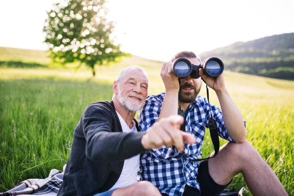 An adult hipster son using binoculars and his senior father sitting on the grass in sunny nature.