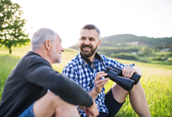 An adult hipster son using binoculars and his senior father sitting on the grass in sunny nature.