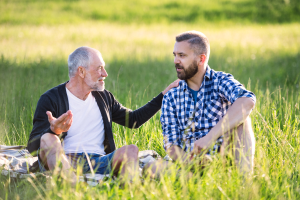 An adult hipster son with his senior father sitting on the grass in sunny nature.