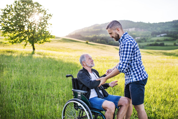 An adult hipster son looking at his senior father in wheelchair on a walk on a meadow in nature at sunset.