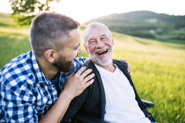 An adult hipster son with his senior father in wheelchair on a walk on a meadow in nature at sunset, laughing. Close up.