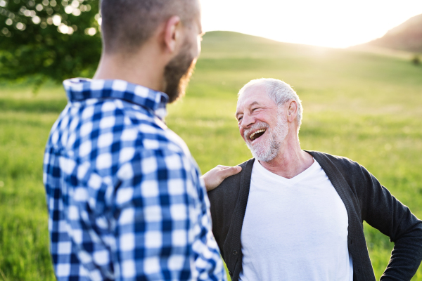 An adult hipster son with his senior father on a walk in nature at sunset.