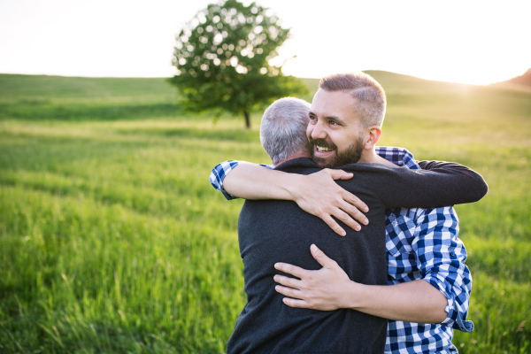 An adult hipster son with his senior father on a walk in nature at sunset, hugging. Copy space.