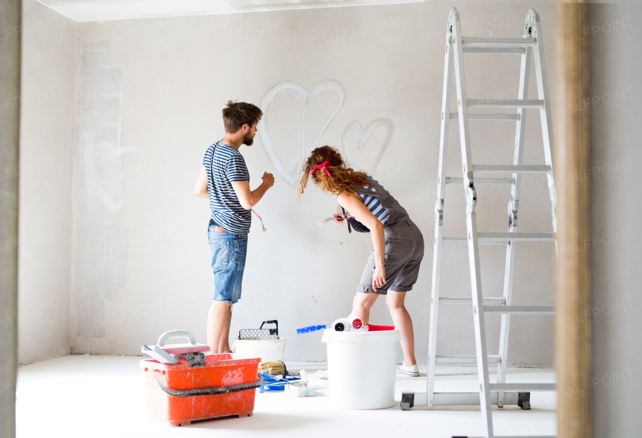 Beautiful young couple having fun and painting walls using paint roller in their new house. Home makeover and renovation concept.