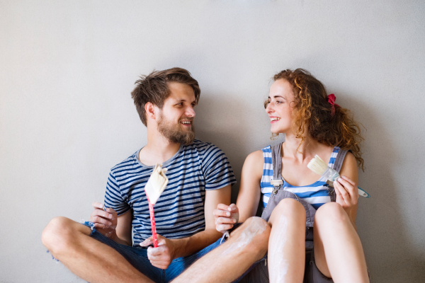 Beautiful young couple in love painting walls of their new house, sitting on the floor, holding paintbrushes. Home makeover and renovation concept.