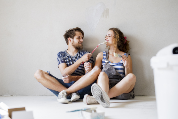 Beautiful young couple in love painting walls of their new house, sitting on the floor, holding paintbrushes. Home makeover and renovation concept.