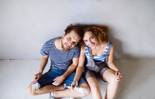 Beautiful young couple in love painting walls of their new house, sitting on the floor, holding paintbrushes. Home makeover and renovation concept.