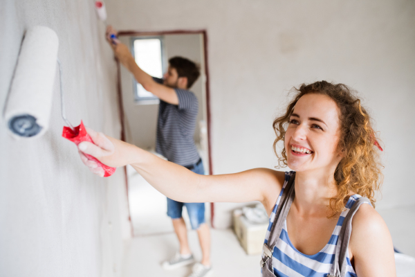 Beautiful young couple having fun and painting walls using paint rollers in their new house. Home makeover and renovation concept.