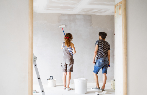 Beautiful young couple having fun and painting walls using paint roller in their new house. Home makeover and renovation concept. Rear view.