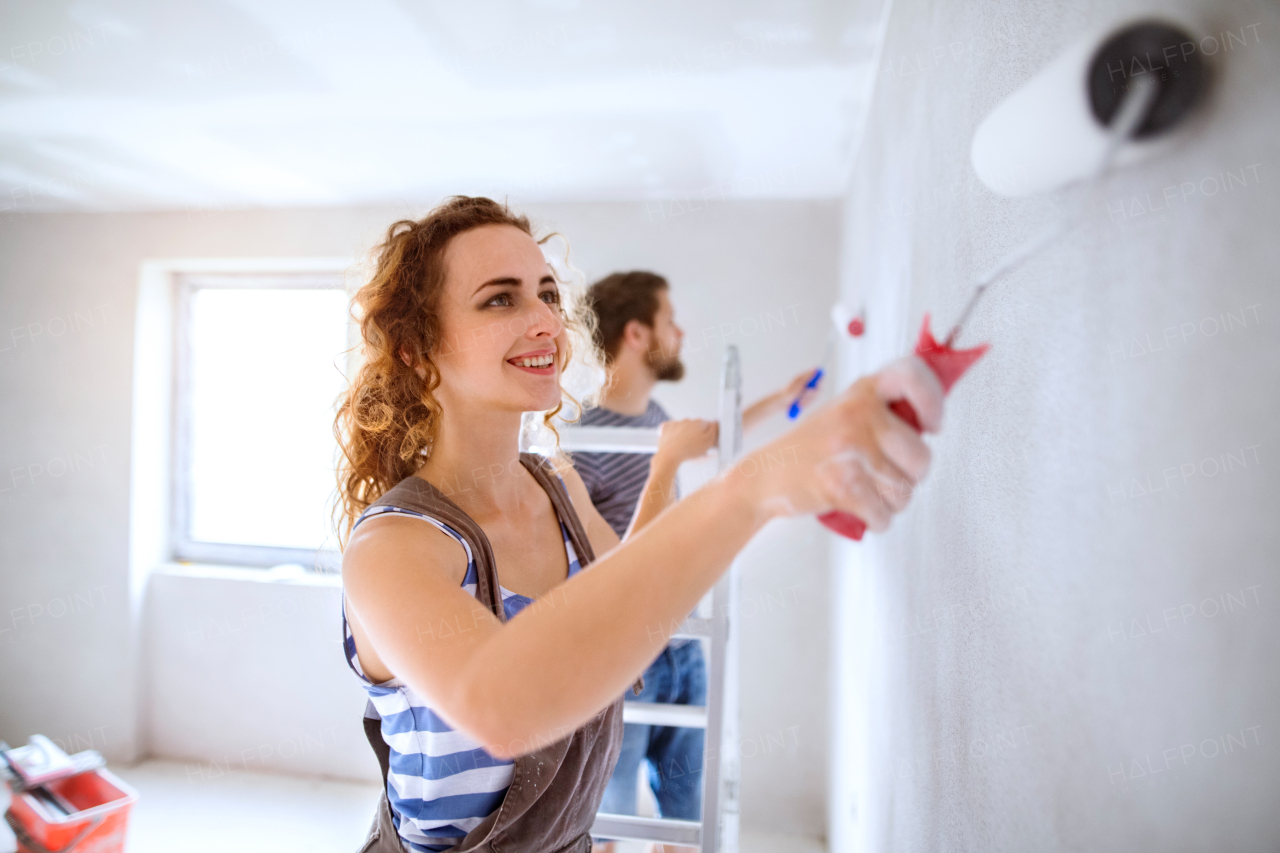 Beautiful young couple having fun and painting walls using paint rollers in their new house. Home makeover and renovation concept.