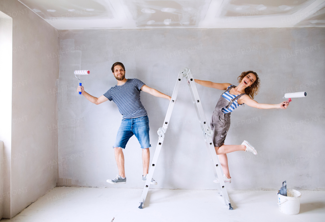 Beautiful young couple at home standing on ladder painting walls in their new house using paint rollers. Home makeover and renovation concept.