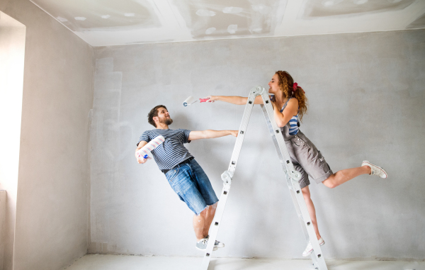 Beautiful young couple at home standing on ladder painting walls in their new house using paint rollers. Home makeover and renovation concept.