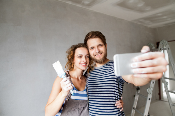 Beautiful young couple in love having fun, painting walls of their new house, taking selfie with smart phone. Home makeover and renovation concept.