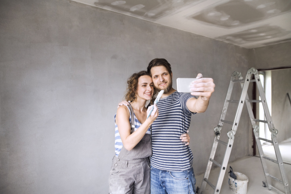 Beautiful young couple in love having fun, painting walls of their new house, taking selfie with smart phone. Home makeover and renovation concept.