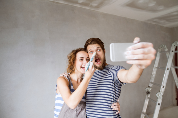 Beautiful young couple in love having fun, painting walls of their new house, taking selfie with smart phone. Home makeover and renovation concept.