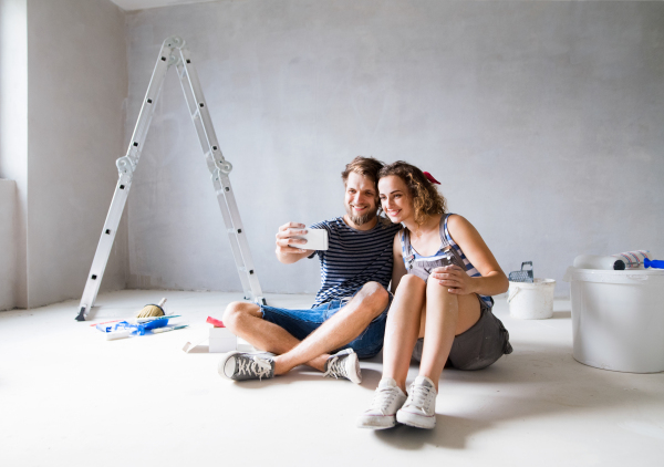 Beautiful young couple in love having fun, painting walls of their new house, taking selfie with smart phone. Home makeover and renovation concept.