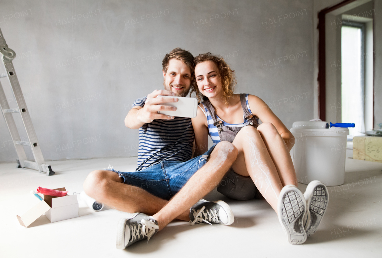 Beautiful young couple in love having fun, painting walls of their new house, taking selfie with smart phone. Home makeover and renovation concept.