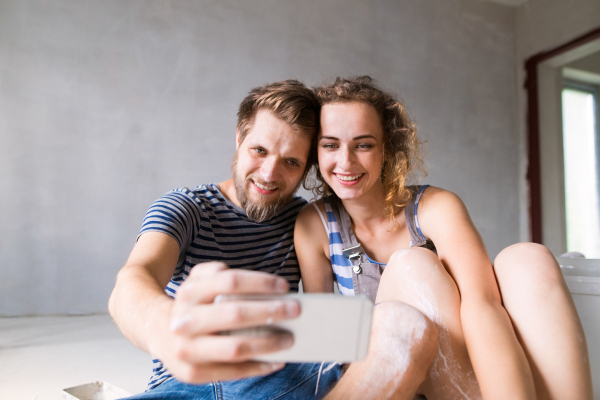 Beautiful young couple in love having fun, painting walls of their new house, taking selfie with smart phone. Home makeover and renovation concept.