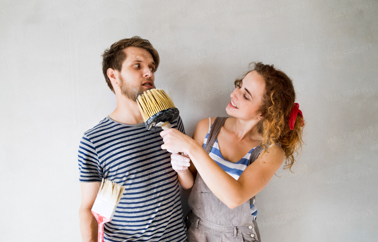 Beautiful young couple in love having fun, painting walls of their new house with paintbrushes, making funny face. Home makeover and renovation concept.