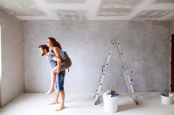 Beautiful young couple having fun and painting walls in their new house. Young man giving his girlfriend piggyback. Home makeover and renovation concept.