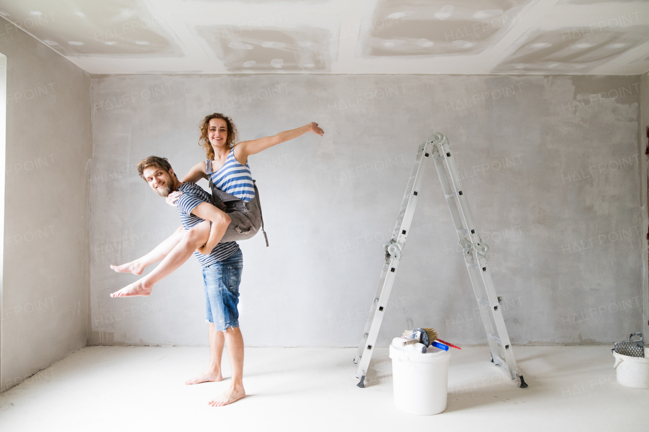 Beautiful young couple having fun and painting walls in their new house. Young man giving his girlfriend piggyback. Home makeover and renovation concept.