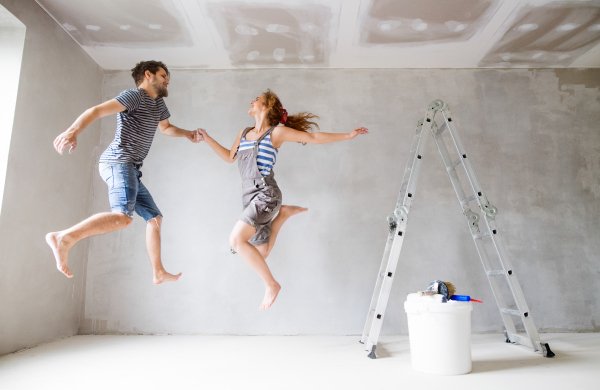 Beautiful young couple having fun and painting walls in their new house, jumping up high. Home makeover and renovation concept.