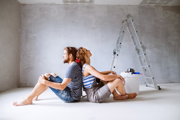 Beautiful young couple painting walls in their new house, sitting on the floor, dreaming. Home makeover and renovation concept.