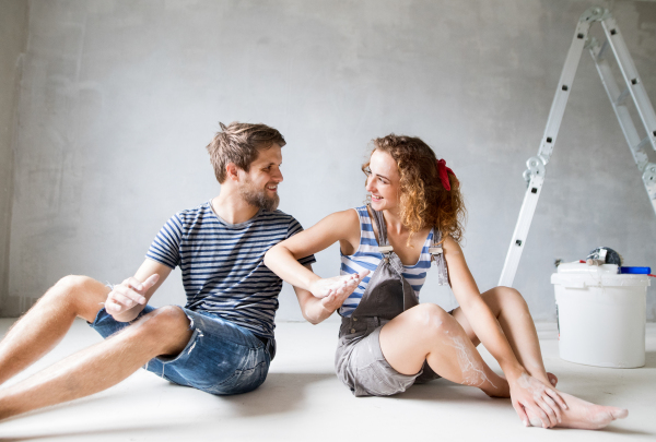 Beautiful young couple painting walls in their new house, sitting on the floor. Home makeover and renovation concept.