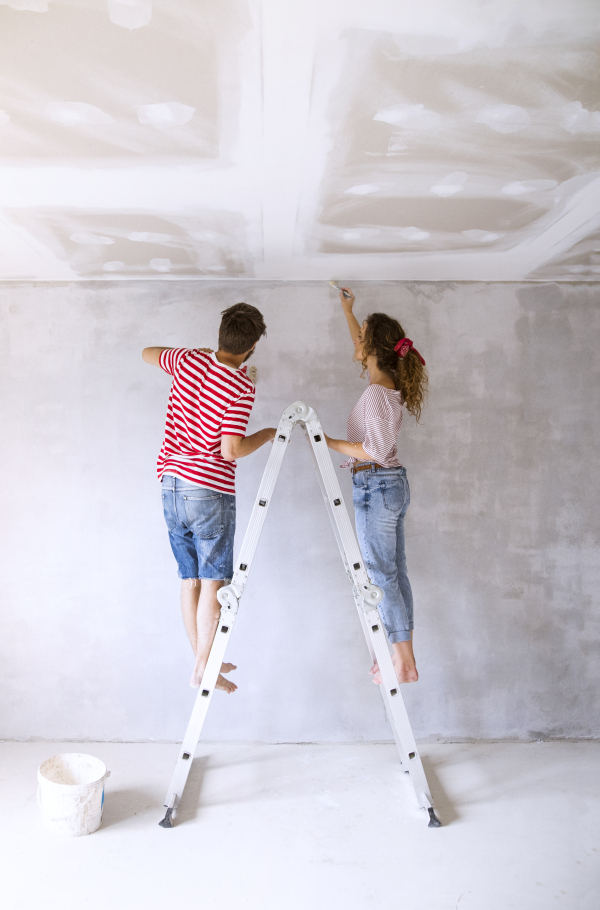 Beautiful young couple standing on ladder painting walls in their new house using paint brushes. Home makeover and renovation concept. Rear view.