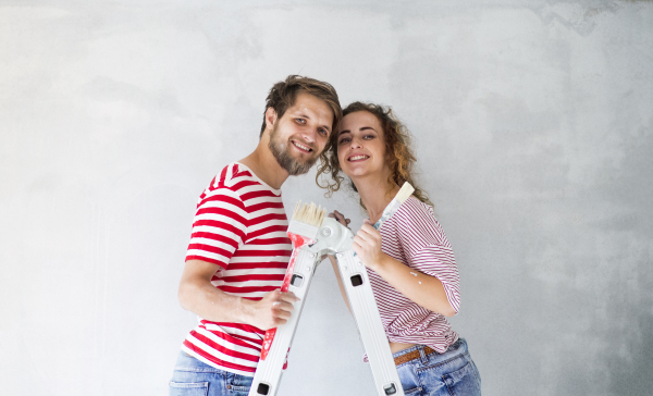 Beautiful young couple standing on ladder painting walls in their new house using paint brushes. Home makeover and renovation concept.