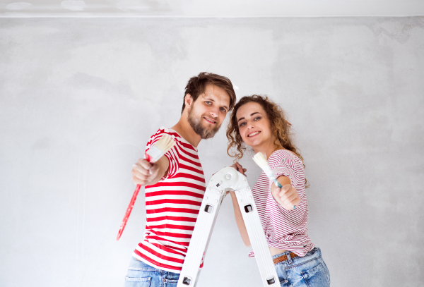 Beautiful young couple standing on ladder painting walls in their new house using paint brushes. Home makeover and renovation concept.