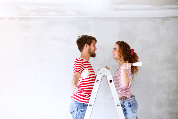 Beautiful young couple standing on ladder painting walls in their new house using paint brushes. Home makeover and renovation concept.