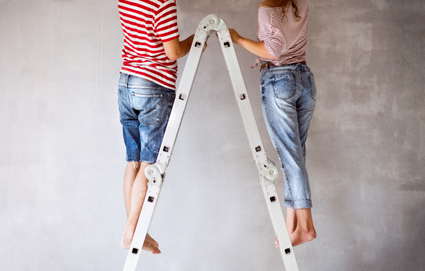 Unrecognizable young couple standing on ladder painting walls in their new house. Home makeover and renovation concept. Rear view.