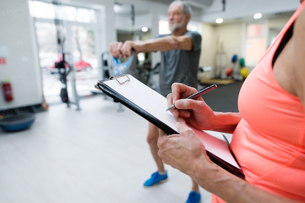 Fit senior man in sports clothing in gym working out with kettlebell. Unrecognizable personal trainer writing his progress on clipboard.