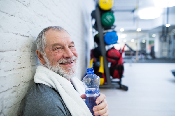 Fit senior man in sports clothing in gym resting after working out, towel around his neck, holding a watter bottle