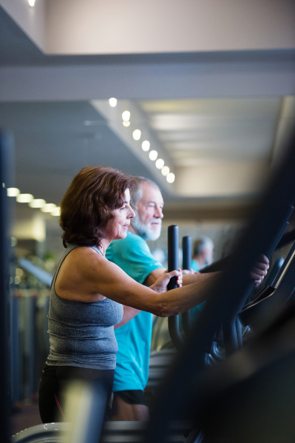 Beautiful fit senior couple in sports clothing in gym doing cardio workout, exercising on elliptical trainer machine. Sport fitness and healthy lifestyle concept.