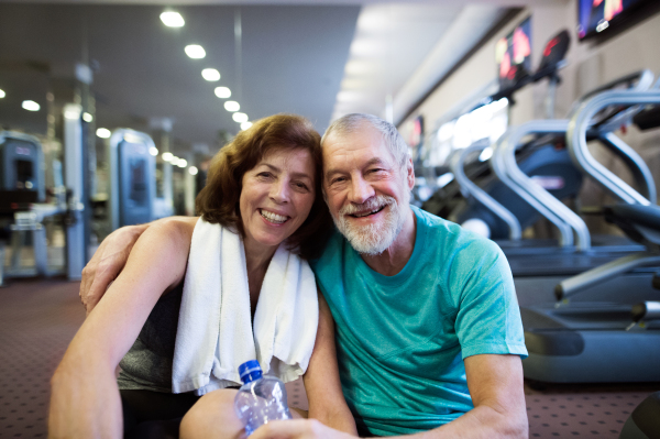 Beautiful fit senior couple in sports clothing in gym resting after working out, sitting in front of treadmills